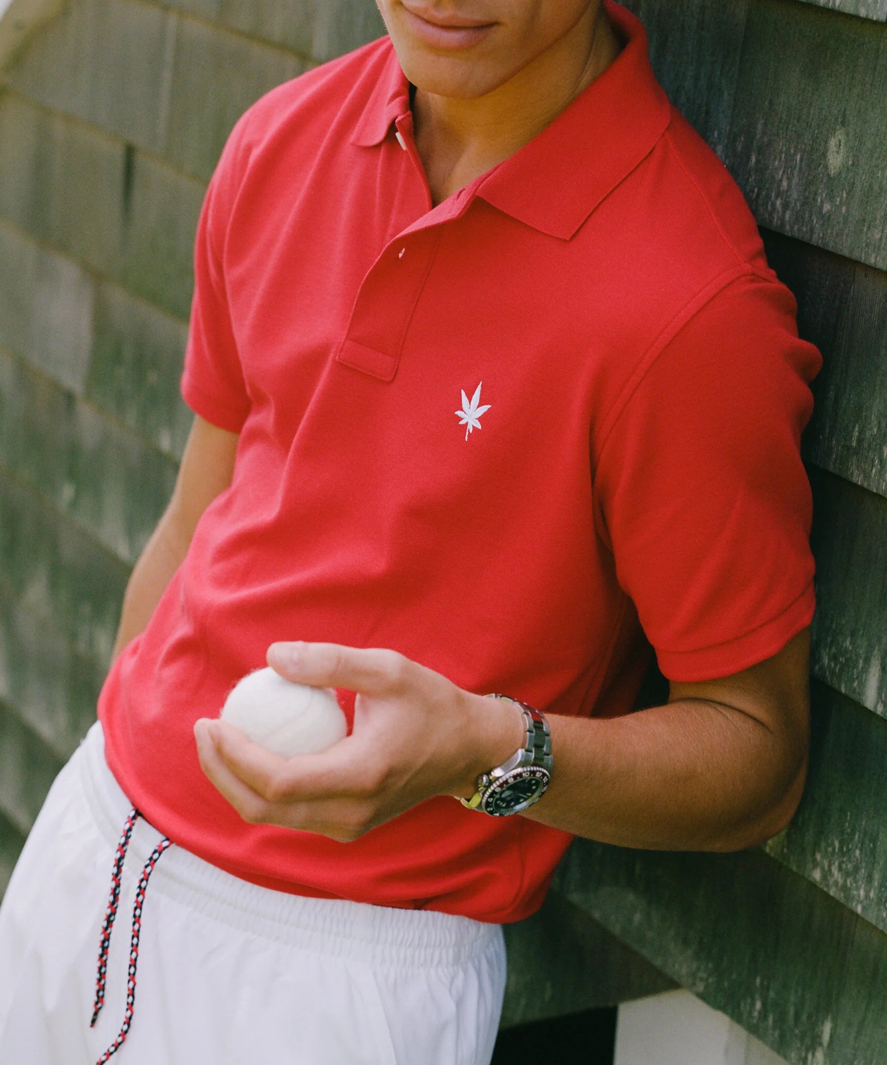 Person wearing a red polo shirt with a logo and white shorts, holding a white ball against a green wall.