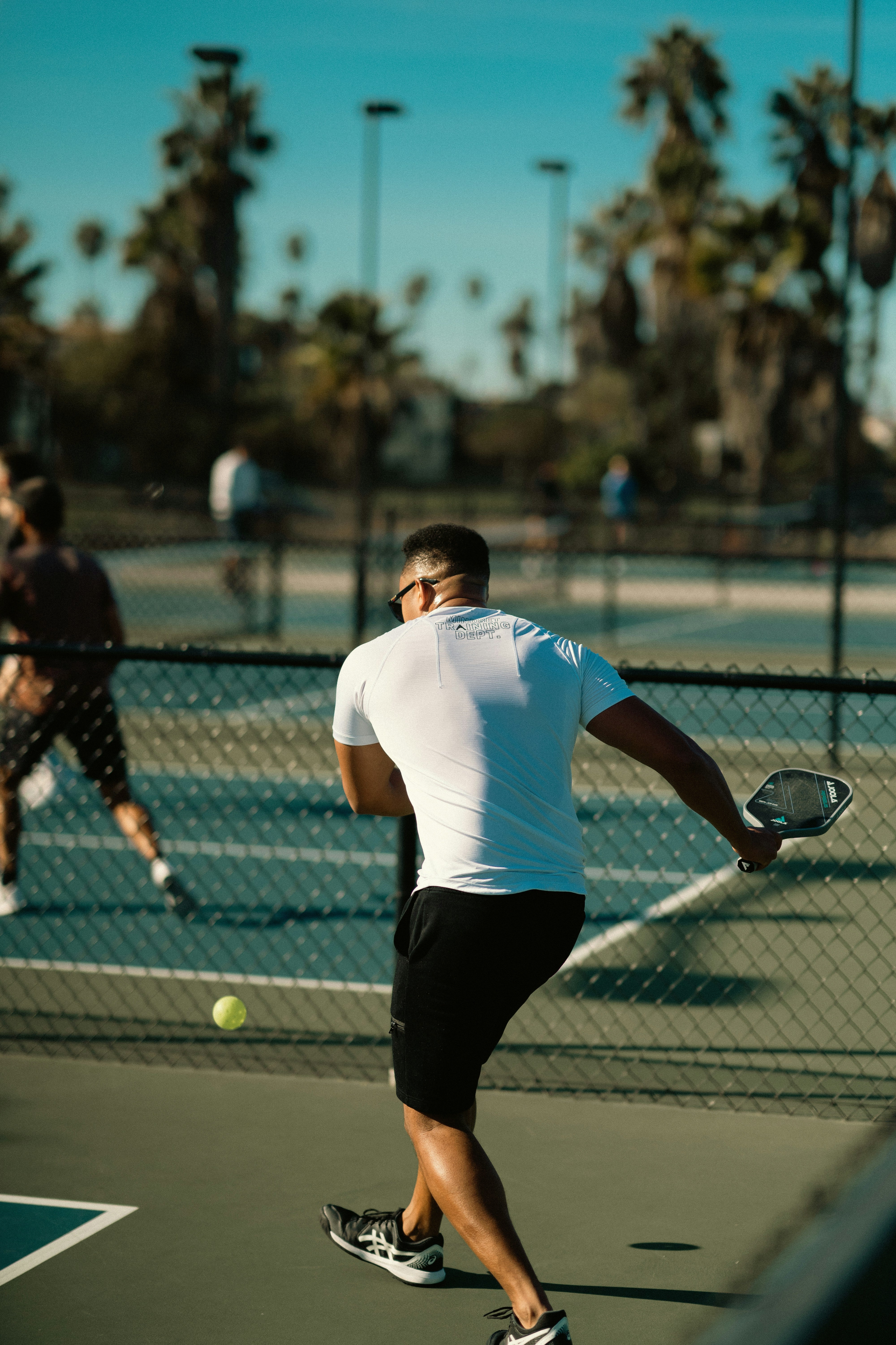 Person playing tennis on an outdoor court with palm trees in the background